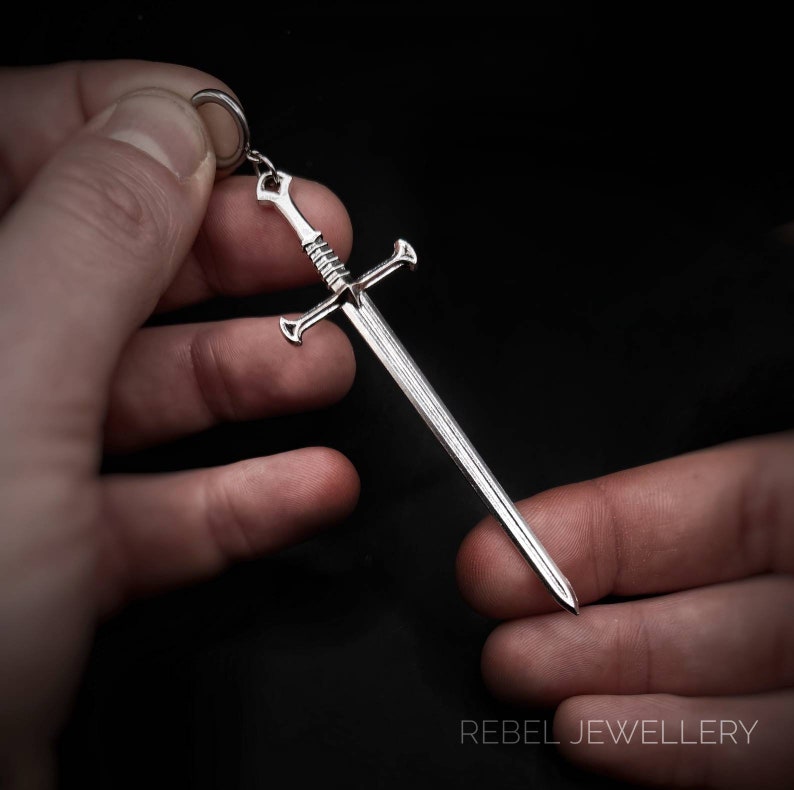 May include: A silver sword pendant with a detailed design. The sword has a pointed tip and a cross guard. The pendant is hanging from a silver chain.
