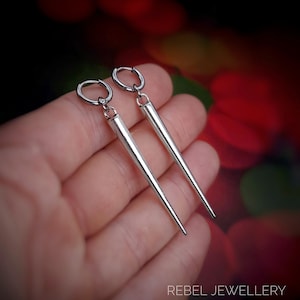 May include: A pair of silver-colored earrings with a long, tapered design. The earrings have a hoop closure and a pointed end. The image is set against a blurred background with the text "REBEL JEWELLERY" at the bottom.