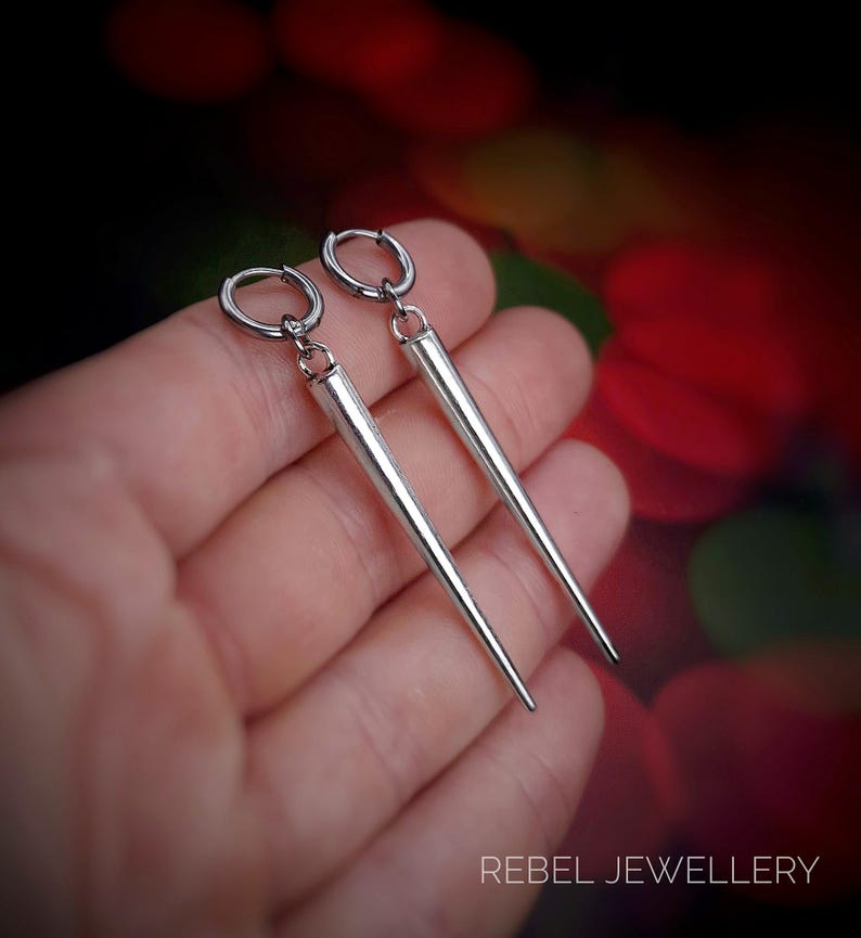 May include: A pair of silver-colored earrings with a long, tapered design. The earrings have a hoop closure and a pointed end. The image is set against a blurred background with the text "REBEL JEWELLERY" at the bottom.
