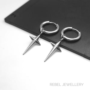 May include: A pair of silver-colored hoop earrings with a star pendant. The earrings are made of a shiny metal and feature a four-pointed star design. The earrings are displayed on a black surface. The text "REBEL JEWELLERY" is at the bottom.