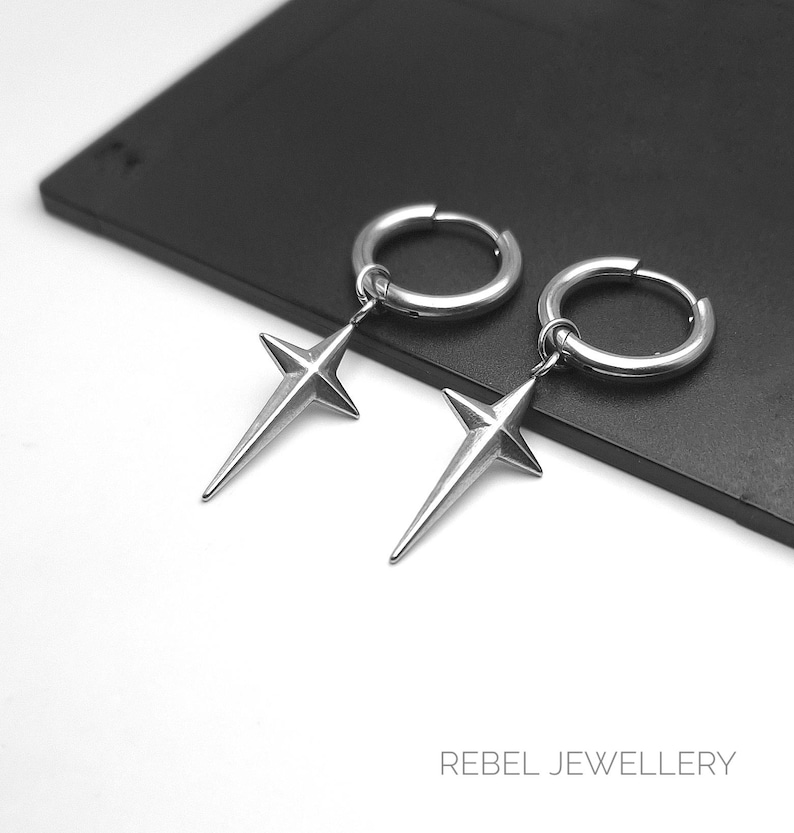 May include: A pair of silver-colored hoop earrings with a star pendant. The earrings are made of a shiny metal and feature a four-pointed star design. The earrings are displayed on a black surface. The text "REBEL JEWELLERY" is at the bottom.