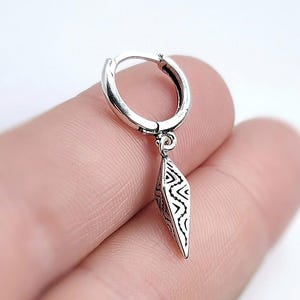 May include: A silver earring with a geometric pendant. The earring is a small hoop with a dangling, diamond-shaped charm. The charm features a detailed, black and white tribal pattern. The earring is shown against a blurred background.