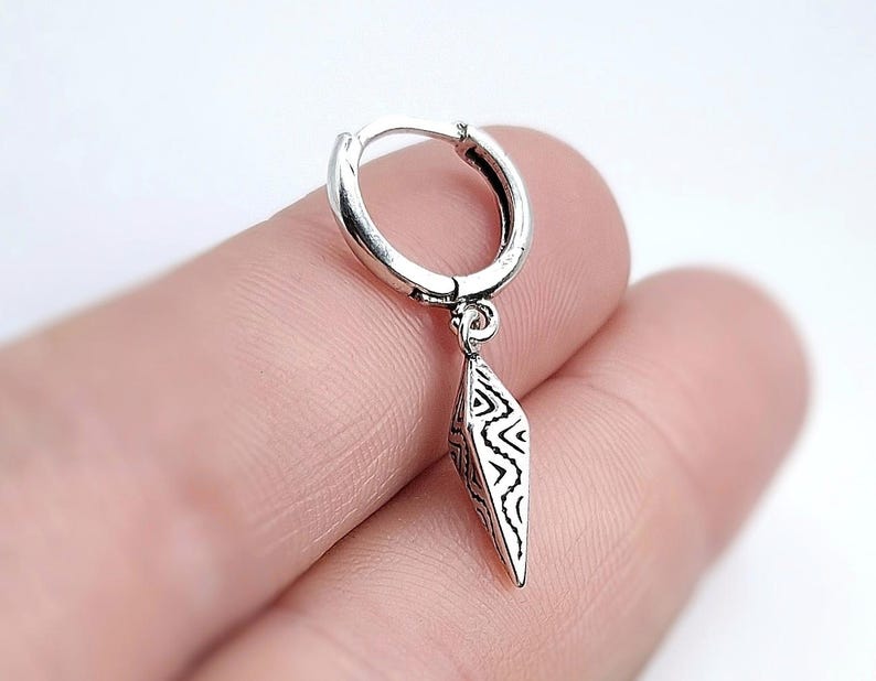 May include: A silver earring with a geometric pendant. The earring is a small hoop with a dangling, diamond-shaped charm. The charm features a detailed, black and white tribal pattern. The earring is shown against a blurred background.