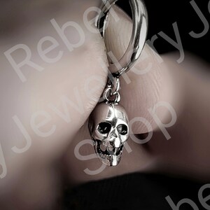 May include: A silver skull earring with a hinged hoop clasp. The skull has detailed facial features, including eye sockets, a nose cavity, and teeth. The earring is held up by a person's fingers.