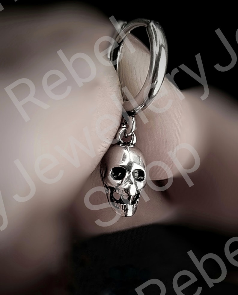 May include: A silver skull earring with a hinged hoop clasp. The skull has detailed facial features, including eye sockets, a nose cavity, and teeth. The earring is held up by a person's fingers.