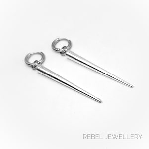 May include: A pair of silver-colored earrings with a hoop closure and a long, conical pendant. The earrings are displayed on a white background. The text "REBEL JEWELLERY" is at the bottom of the image.