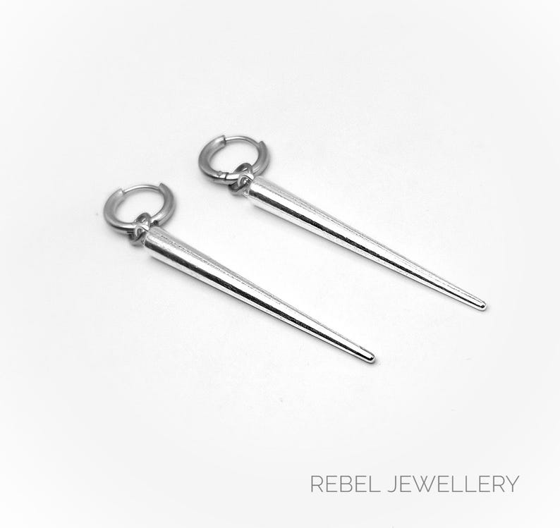 May include: A pair of silver-colored earrings with a hoop closure and a long, conical pendant. The earrings are displayed on a white background. The text "REBEL JEWELLERY" is at the bottom of the image.