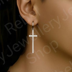 May include: A silver-colored cross earring with a hoop closure. The cross is a simple, elongated design, hanging from the ear. The earring is shown against a blurred background.