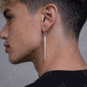 May include: A silver, linear earring with a long, tapered design. The earring is suspended from a small hoop and hangs vertically. The jewelry is shown on a person's ear, with a black shirt in the background.