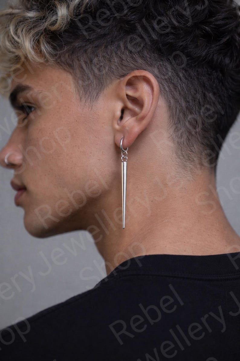 May include: A silver, linear earring with a long, tapered design. The earring is suspended from a small hoop and hangs vertically. The jewelry is shown on a person's ear, with a black shirt in the background.