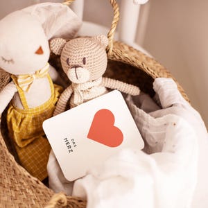 May include: A white card with a red heart and the text "DAS HERZ" in black. The card is in a wicker basket with a crocheted bunny and bear.
