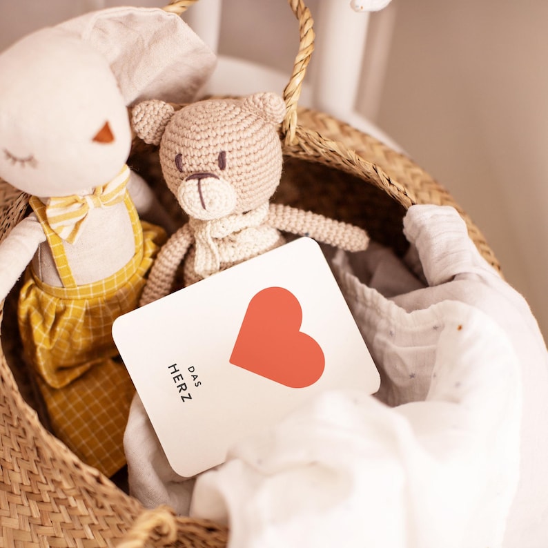 May include: A white card with a red heart and the text "DAS HERZ" in black. The card is in a wicker basket with a crocheted bunny and bear.