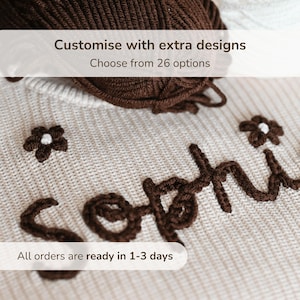 Può includere: Primo piano di un tessuto a maglia beige con il nome "Sophie" ricamato in filo marrone. Il design include piccoli accenti floreali. Il testo "Personalizza con design extra" e "Scegli tra 26 opzioni" è visibile.