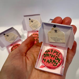 May include: Miniature pink cake with green frosting reading "Happee Birthdae Harry" in a white box. The box has a small tag attached. Several similar boxes are visible.