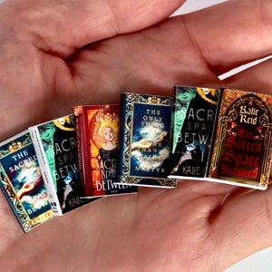 May include: A selection of small books with vibrant covers, held in a hand. Titles visible include "The Sacred Space Between" and "The Only One Than Martyr". The books feature gold and blue detailing, with various illustrations.