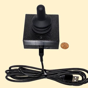 Könnte beinhalten: Schwarzer Joystick-Controller mit abgerundeter Oberseite und quadratischer Basis. Ein USB-Kabel ist mit der Basis verbunden. Eine Münze liegt neben dem Controller zum Größenvergleich. Der Controller befindet sich auf einem hellgelben Hintergrund.