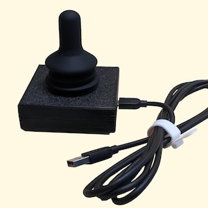 Könnte beinhalten: Ein schwarzer USB-Joystick mit einem runden, schwarzen Steuerknüppel und einem schwarzen Sockel. Der Joystick ist mit einem schwarzen USB-Kabel verbunden.