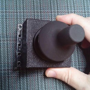 May include: A black, square-shaped electronic component featuring a textured finish and a prominent, cylindrical knob extending from the center. The base includes a side flange with five small, circular mounting holes for secure installation.
