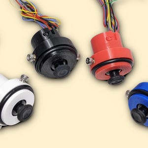 Op de afbeelding: Vier elektronische componenten in wit, zwart, rood en blauw. Elke component heeft een zwarte lens, een cirkelvormig ontwerp en een bundel kleurrijke draden. De componenten zijn gerangschikt op een lichtgele achtergrond.