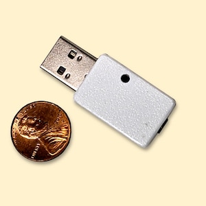 May include: A small, silver USB flash drive with a metallic connector is positioned next to a copper penny. The flash drive has a rectangular body with a small black dot and a black component on the side. The penny is a US one-cent coin.