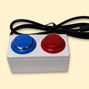 May include: A white rectangular device with two large, round buttons, one blue and one red. A black cable with a 3.5mm jack extends from the device. The buttons are raised and appear to be made of plastic.
