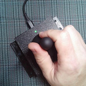May include: A black, textured, rectangular joystick controller featuring a central spherical handle, a single green LED indicator light, and a black braided USB cable connected to the top edge of the device housing.