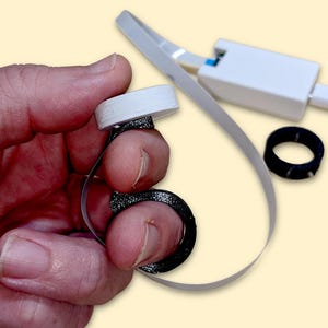 May include: A white and black finger-worn device with a connected white cable and a separate black ring. The device is held by a hand. The device appears to be a wearable tech product.