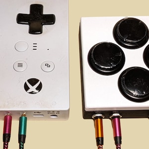 Puede incluir: Una consola de juegos blanca con cuatro botones negros y una almohadilla circular negra en la parte superior. La consola tiene un logotipo de Xbox negro y cuatro cables de colores conectados a la parte inferior.