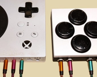 May include: A white Xbox Adaptive Controller featuring a black D-pad, menu buttons, and an Xbox logo, positioned next to a square white auxiliary button interface with four large black circular buttons, both connected to colourful braided cables.