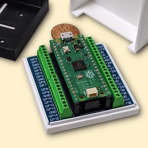 May include: A green Raspberry Pi Pico H 2021 circuit board with green terminal blocks is mounted on a white base. A black box and a white box are in the background. The board has the text "Raspberry Pi Pico H 2021".