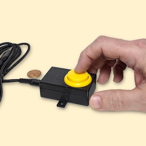 May include: A black electronic device with a yellow button is being pressed by a hand. The device has a black cord and a small coin is next to it. The background is a light yellow color.