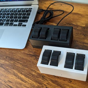 Op de afbeelding: Twee rechthoekige macro-toetsenbordcontrollers, één in zwart en één in wit, elk voorzien van vier zwarte geribbelde scrollwielen. De controllers zijn verbonden met zwarte kabels en staan naast een zilveren laptop op een houten ondergrond.