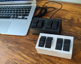 May include: Two rectangular macro keypad controllers, one in black and one in white, each featuring four black ridged scroll wheels. The controllers are connected by black cables and positioned next to a silver laptop on a wood grain surface.