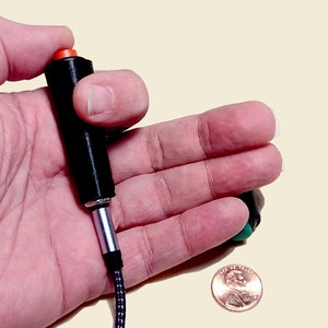 Può includere: Un dispositivo nero e argento con un pulsante arancione è tenuto in mano. Il dispositivo ha un cavo collegato. Una moneta è accanto al dispositivo.