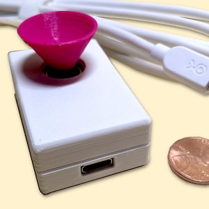 Puede incluir: Un dispositivo rectangular blanco con un embudo rosa en la parte superior. El dispositivo tiene un puerto USB-C en el lateral y un cable blanco conectado. Una moneda de un centavo se coloca junto al dispositivo para la escala.