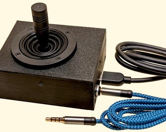 May include: A black joystick controller with a circular base and a cylindrical handle. It has a USB cable, a 3.5mm audio jack, and a blue braided cable. The controller is designed for gaming or other interactive applications.