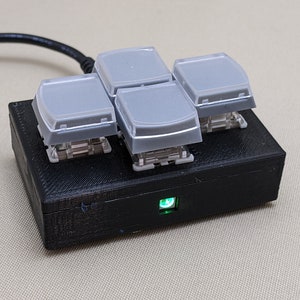 May include: A black 3D printed box with four gray buttons and a green LED light. The box is connected to a black cable.