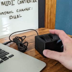 Könnte beinhalten: Ein schwarzer, rechteckiger MIDI-Controller mit zwei texturierten Drehrädern auf der Oberseite. Das Gerät ist über ein schwarzes Kabel mit einem silbernen Laptop verbunden. Der Text SB MIDI CONTROLLER und CONTROL CHANGE: DUAL ist auf einer Tafel.