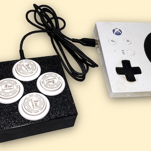 Könnte beinhalten: Ein weißer Xbox-Controller ist mit einer schwarzen Box mit vier weißen, kreisförmigen Knöpfen verbunden. Der Controller hat ein schwarzes Steuerkreuz und eine schwarze kreisförmige Taste. Ein schwarzes Kabel verbindet die beiden Geräte.