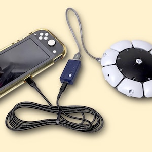 May include: A handheld gaming console in a clear protective case connected via a black cable and a small blue adapter to a circular, white and black accessibility controller. The controller features a central plus sign and numbered buttons.