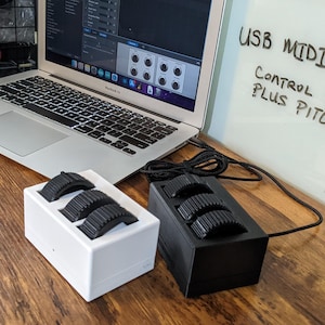 May include: A white and a black rectangular USB MIDI controller, each featuring three black ridged control wheels. The devices are connected by a black cable and positioned next to a laptop displaying audio software and a sign reading USB MIDI CONTROLLER.