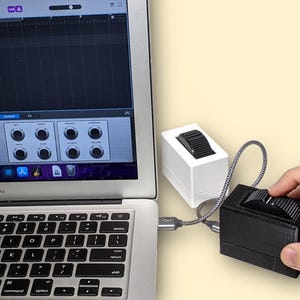 Può includere: Un computer portatile argentato che mostra un'interfaccia di workstation audio digitale, collegato tramite un cavo intrecciato a un controller rettangolare nero con una rotellina testurizzata, accanto a un'unità bianca identica.
