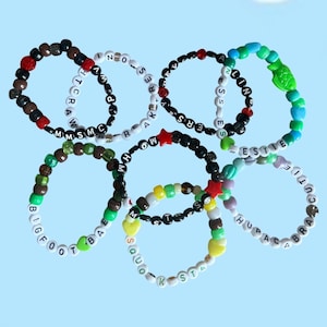 Peut inclure: Plusieurs bracelets de perles colorées, chacun épelant des mots et des phrases avec des perles de lettres noires. Les bracelets présentent diverses couleurs, notamment le rouge, le vert, le jaune et le bleu, sur un fond bleu clair.