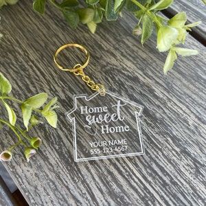 May include: A clear acrylic keychain shaped like a house with the text "Home Sweet Home" and a space for a name and phone number. The keychain has a gold metal ring.
