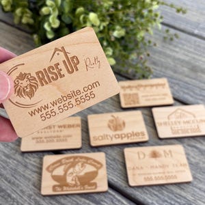 May include: A collection of wooden business cards with various logos and contact information. The cards are engraved with text, including "Rise Up Realty", "Saltyapples", "Wesley Mecune", and "DM Dana Mandy Team".