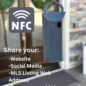 May include: A gray keychain with a small black NFC chip. The keychain has a loop for attaching to a key ring. The text "Share your: -Website -Social Media -MLS Listing Web Address -Reprogrammable!" is visible below the keychain.