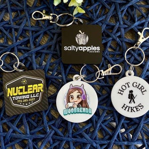 May include: Four different keychains with various designs. The first keychain is black with a white logo that says "Nuclear Towing LLC" and a phone number. The second keychain is black with a white apple logo and the text "saltyapples designs". The third keychain is white with a cartoon image of a girl wearing headphones and the text "WooBrenda". The fourth keychain is white with the text "Hot Girl Hikes" and a silhouette of a person hiking.