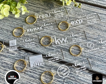 Może przedstawiać: Kolekcja prostokątnych breloków z przezroczystego akrylu z różnymi logotypami firm nieruchomości, napisem home, Your Name oraz 555-123-4567. Każdy brelok jest przymocowany do złotego metalowego kółka na ciemnej drewnianej powierzchni.