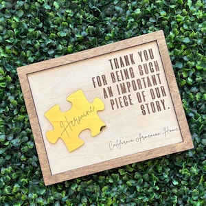 May include: A rectangular wooden sign with a light wood center panel featuring the dark text: THANK YOU FOR BEING SUCH AN IMPORTANT PIECE OF OUR STORY. A gold mirror-finish puzzle piece is attached to the left with the name Hermine in script.