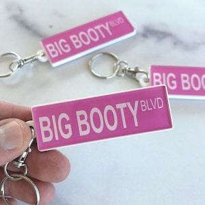 May include: Three rectangular keychains featuring a bright pink background with white text that reads BIG BOOTY BLVD. Each keychain is attached to a silver metal split ring and a lobster claw clasp, providing a secure way to attach keys or accessories.